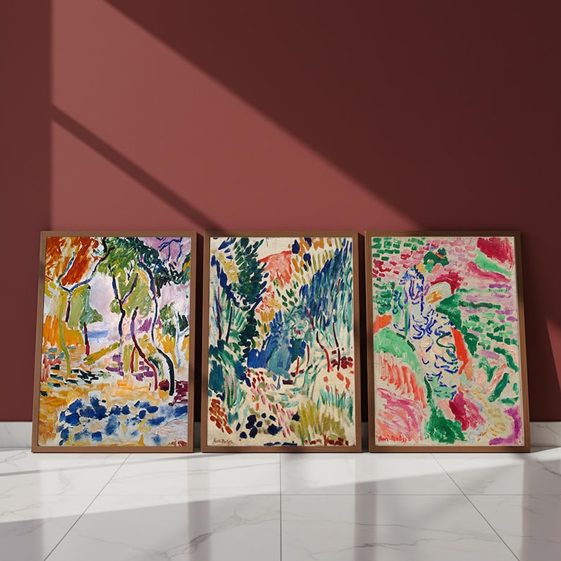 May include: Three framed abstract landscape paintings. The artwork features vibrant colors, including green, blue, orange, and pink. The paintings are in wooden frames and lean against a maroon wall on a white tiled floor.