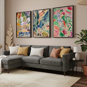 May include: Three framed abstract art prints in vibrant colors hang above a gray sectional sofa with several throw pillows. The room features a neutral color palette with a rug and a side table with a lamp.