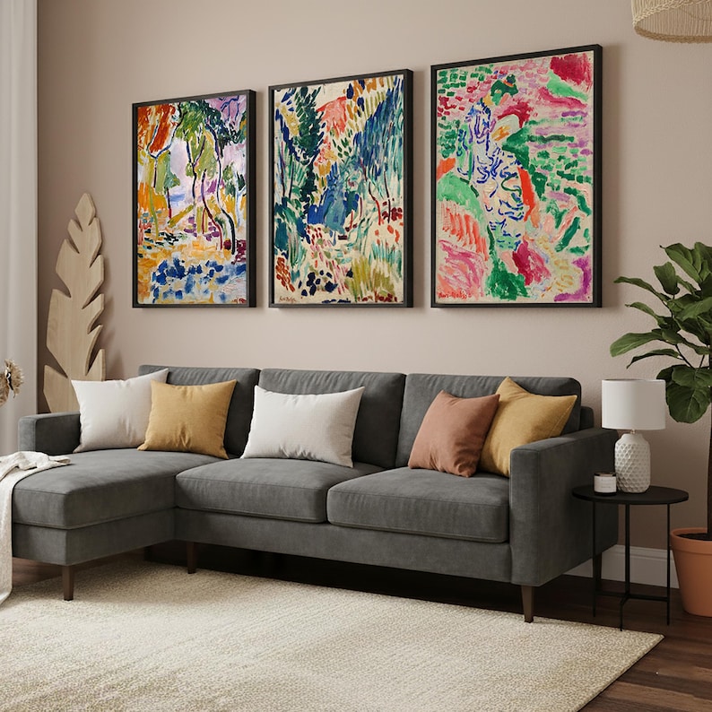 May include: Three framed abstract art prints in vibrant colors hang above a gray sectional sofa with several throw pillows. The room features a neutral color palette with a rug and a side table with a lamp.