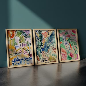 May include: Three framed landscape paintings with colorful abstract art. The artwork features a variety of colors, including green, blue, orange, and pink. The frames are a light wood color, and the paintings are leaning against a teal wall.