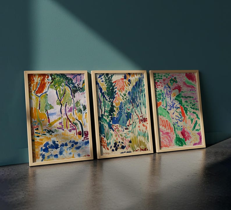 May include: Three framed landscape paintings with colorful abstract art. The artwork features a variety of colors, including green, blue, orange, and pink. The frames are a light wood color, and the paintings are leaning against a teal wall.