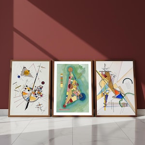 May include: Three framed abstract art prints. Each print features geometric shapes and lines in a variety of colors, including yellow, blue, red, and black. The frames are a warm brown color, and the artwork is displayed against a maroon wall.