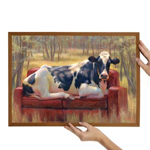 Peut inclure: Une impression d'art encadrée représentant une vache noire et blanche se prélassant sur un canapé rouge dans un cadre forestier. La vache est le point central, avec sa langue sortie. L'œuvre est tenue par deux paires de mains.