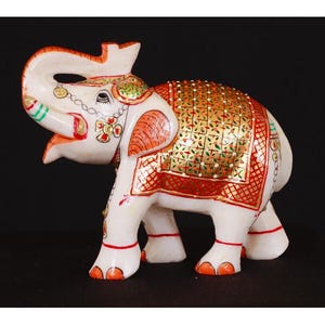 May include: A white elephant ornament with orange and gold detailing. The elephant features a decorative saddle blanket with a gold geometric pattern and red trim. The trunk is raised, and the ears are adorned with green and gold details.
