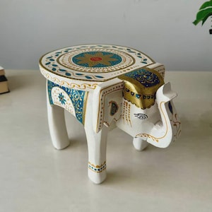 Puede incluir: Un taburete blanco pintado a mano con forma de elefante y parte superior redonda. El taburete presenta intrincados diseños florales azules y dorados en la parte superior y los lados. La cabeza y la trompa del elefante están detalladas con detalles dorados.