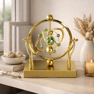 May include: A gold-colored decorative atomic model with a green glass sphere at the center and smaller green spheres. The model is mounted on a rectangular base. The background includes a vase with dried flowers and a lit candle.