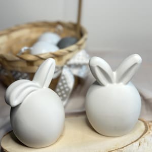 May include: Two white ceramic bunny ornaments with rounded bodies and upright ears, displayed on a wooden slice. A wicker basket with eggs is in the background, suggesting an Easter theme. The bunnies have a smooth, minimalist design.