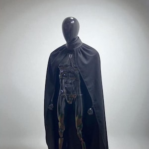 May include: A long, solid black cape with a high collar draped over a glossy black mannequin. The cape features a smooth, matte fabric texture that extends from the shoulders down to the floor, creating a full-length silhouette.