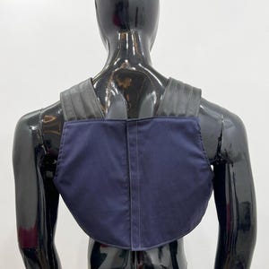 May include: A dark blue chest piece or harness featuring a solid navy fabric front panel with a vertical center seam and wide, black, ribbed shoulder straps that extend over the upper back area, worn on a glossy black mannequin torso.