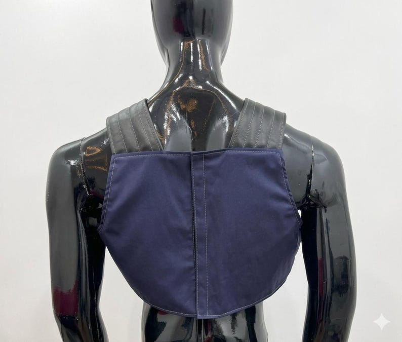 May include: A dark blue chest piece or harness featuring a solid navy fabric front panel with a vertical center seam and wide, black, ribbed shoulder straps that extend over the upper back area, worn on a glossy black mannequin torso.