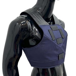 May include: A dark blue chest harness featuring wide, dark gray shoulder straps and multiple black rectangular hook-and-loop fastener patches on the front panel. The garment is displayed on a glossy black mannequin torso against a white background.
