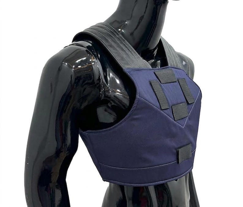 May include: A dark blue chest harness featuring wide, dark gray shoulder straps and multiple black rectangular hook-and-loop fastener patches on the front panel. The garment is displayed on a glossy black mannequin torso against a white background.