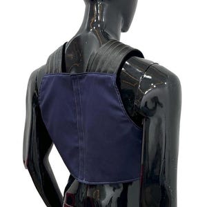 May include: A navy blue vest featuring a structured design with wide black shoulder straps. The garment is constructed with visible vertical stitching along the center back panel and across the shoulder straps, providing a clean, tailored appearance.