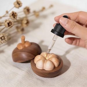 May include: A wooden essential oil diffuser in the shape of a mangosteen fruit. The diffuser is brown with a light-colored interior. A black dropper is held above the diffuser, dispensing liquid. The diffuser is on a white surface.