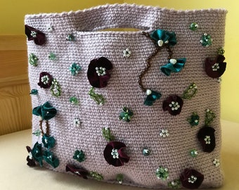 Puede incluir: Una bolsa de mano de crochet lavanda con un diseño de cuentas. La bolsa está decorada con flores de tela color burdeos y turquesa y cuentas.