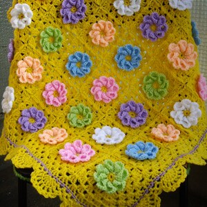 May include: A yellow crocheted baby blanket with a scalloped edge and a purple bow. The blanket is decorated with colorful crocheted flowers in various shades of pink, blue, green, and white.