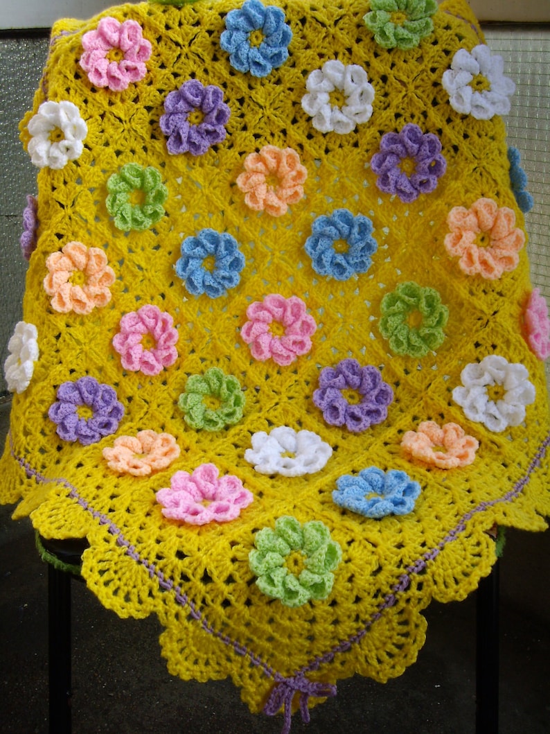 May include: A yellow crocheted baby blanket with a scalloped edge and a purple bow. The blanket is decorated with colorful crocheted flowers in various shades of pink, blue, green, and white.