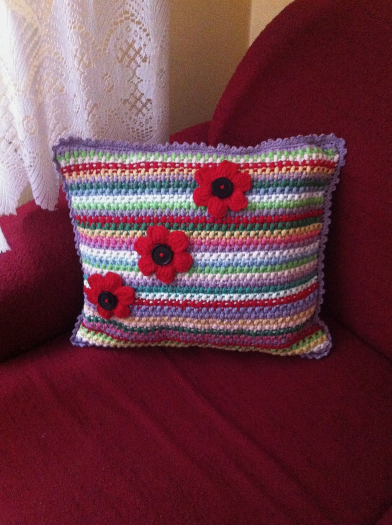 May include: A colorful striped crocheted pillow with three red fabric flowers with black button centers.