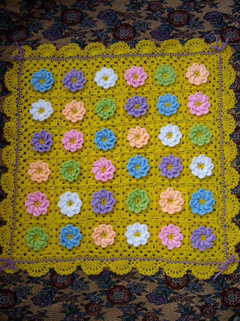 May include: A yellow crocheted baby blanket with a scalloped border. The blanket is made up of squares with different colored flowers in each square. The flowers are in shades of pink, purple, blue, green, and white.