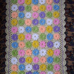 May include: A crocheted baby blanket with a light yellow background and a pattern of colorful flowers. The blanket has a white crocheted border with a scalloped edge.