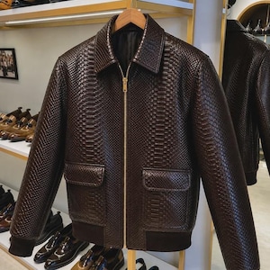 May include: A dark brown jacket with a textured snakeskin pattern, featuring a classic collar, a central gold-toned zip closure, and two front flap pockets. The garment includes dark brown ribbed cuffs and a matching ribbed hem at the waist.