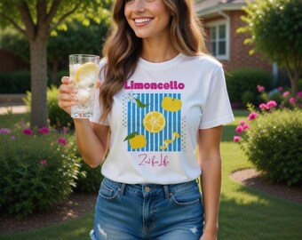 May include: White t-shirt with a "Limoncello" graphic featuring lemons and the text "Zeal for Life." The shirt is paired with denim shorts. The image is set outdoors with a person holding a glass of lemonade.