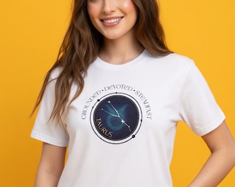 May include: White t-shirt featuring a Taurus zodiac design. The design includes the word "TAURUS" and the words "GROUNDED, DEVOTED, STEADFAST" around a constellation graphic in shades of blue and green.