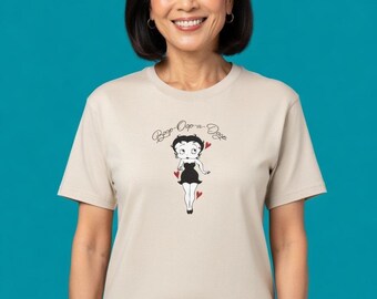 May include: Beige t-shirt featuring a black and white cartoon graphic of a woman with the text "Boop-Oop-a-Doop". The cartoon woman is wearing a black dress and has red heart accents. The t-shirt is short-sleeved.