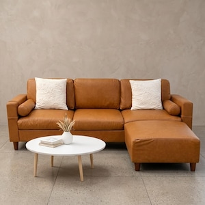 May include: A tan leather sectional sofa with a chaise lounge, featuring two white square throw pillows with embossed patterns and two cylindrical bolster pillows. A round white coffee table with light wood legs sits in front.