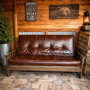 May include: A brown leather futon sofa with a wooden frame. The sofa has a tufted design and is placed in a rustic setting with wooden walls and a tiled floor. A sign that says "CABIN RULES" is visible.