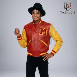 May include: A red and yellow varsity jacket featuring a large letter M on the left chest, snap button closure, and ribbed cuffs. The jacket is worn with a black fedora hat and black trousers. The TASHION logo is visible in the top right corner.