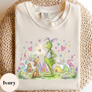 May include: Ivory-colored t-shirt featuring a whimsical Easter-themed illustration. The design showcases a green character and a dog, both wearing bunny ears, set against a colorful village backdrop with hearts and Easter eggs. The shirt has a crew neck and short sleeves.