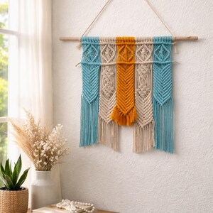 May include: A macrame wall hanging with a wooden dowel. The design features sections of turquoise, beige, and mustard yellow knotted cotton cord. The wall decor is suspended by a natural fiber cord.