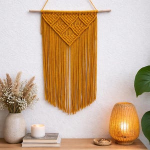 May include: Mustard yellow macrame wall hanging with a geometric design and fringe. The wall decor is suspended from a wooden dowel. A wooden shelf below displays a vase of dried flowers, books, a candle, and a woven lamp.