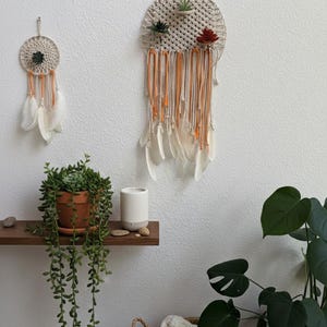 May include: Two macrame dream catchers with feathers and orange accents hang on a white wall. A small potted plant and a decorative shelf with a white candle holder are also visible. A basket with a blanket and a large potted plant complete the scene.