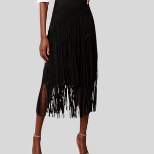 May include: A black midi-length skirt featuring a high-waisted design and multiple layers of long, thin fringe that hang from the waist down to the hem. The skirt is paired with a crisp white button-down shirt and simple strappy sandals.