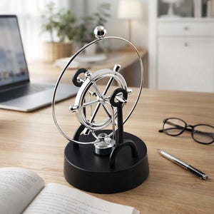 Op de afbeelding: Een zilverkleurig Newton's cradle kinetisch kunst bureau speelgoed met een zwarte basis. Het speelgoed heeft een wielachtig ontwerp met een zilveren bal aan de bovenkant. Een laptop, bril, pen en open boek staan ook op het houten bureau.