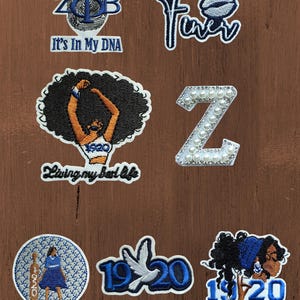 Peut inclure: Un ensemble de sept écussons brodés aux motifs bleus, blancs et noirs. Les éléments incluent le texte ZPhi, It's In My DNA, Finer, Living my best life, 1920, une lettre Z stylisée avec des perles, une colombe et des illustrations de personnages.