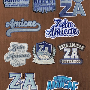 May include: A collection of ten embroidered patches in shades of blue and white. The patches feature text including AMICAE, ZETA AMICAE, I HEART ZA, and SISTERHOOD, along with imagery of hands shaking, a shield, and a heart, all celebrating Zeta Amicae.