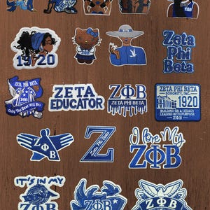 Peut inclure: Une collection de vingt écussons brodés présentant des motifs bleus et blancs liés à la sororité Zeta Phi Beta. Les écussons incluent diverses illustrations de personnages, les lettres grecques ZΦB, l'année 1920 et du texte comme Zeta Educator.