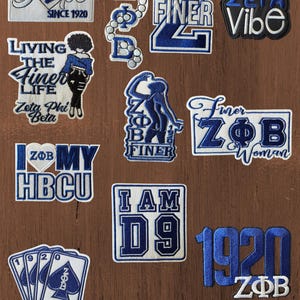 May include: A collection of embroidered patches featuring blue and white designs related to Zeta Phi Beta. The patches include text such as Unapologetically Dope, Living The Finer Life, Zeta Vibe, I Am D9, and 1920, along with various symbols like a dove, playing cards, and Greek letters.