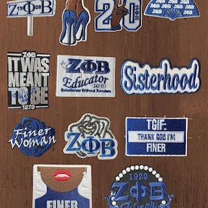 May include: A collection of twelve embroidered patches featuring blue and white designs, including text such as Finer Way, ZPhiB, Nineteen 20, It Was Meant To Be, Educator, Sisterhood, Finer Woman, TGIF, and Zeta Phi Beta, alongside graphic icons.