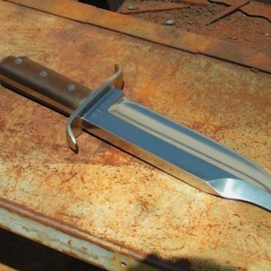 May include: A fixed-blade knife featuring a polished silver-tone steel blade with a prominent central fuller and a curved edge. The handle is constructed from smooth brown material secured with three metal rivets and includes a metal crossguard.