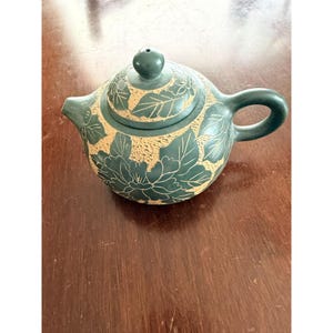 May include: A small, dark green ceramic teapot with a lid and handle. The teapot features a floral design with etched leaves and flowers in a lighter tan color. The teapot is sitting on a dark brown wooden surface.