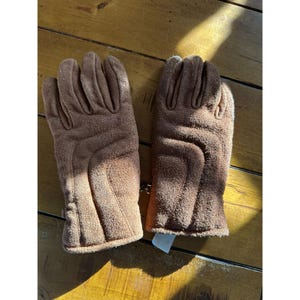 May include: A pair of brown gloves made of a soft, suede-like material. The gloves have a classic design with individual finger slots and a textured surface. The gloves are displayed on a wooden surface.