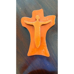 May include: Wooden wall art featuring a stylized figure with outstretched arms, layered over a cross-shaped background. The figure and cross are made of different shades of wood, with an orange background. The art piece is likely handmade.