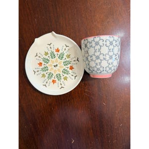 May include: A white ceramic spoon rest and a matching cup. The spoon rest features a leaf design in green, orange, and yellow. The cup has a floral pattern in blue and gray with a pink base. The items are on a dark wood surface.
