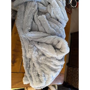 May include: A light gray, plush robe with a soft, fuzzy texture. The robe is folded on a wooden surface, showcasing its thick, comfortable material. The robe appears to be made of a warm, cozy fabric, perfect for lounging.