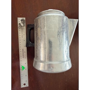 May include: A vintage aluminum coffee pot with a black handle and a spout. The pot has a cylindrical body with a lid and is approximately 10 inches tall. A stainless steel ruler is next to the pot.