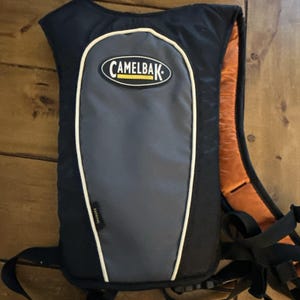 May include: A black and gray CamelBak hydration pack with orange shoulder straps. The pack features a gray center panel, a white outline, and the CamelBak logo. Black straps and a small tag are also visible.
